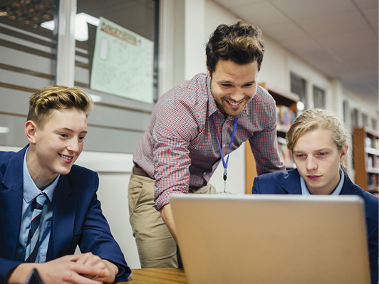 Two students in high school using a computer. A teacher is helping them.